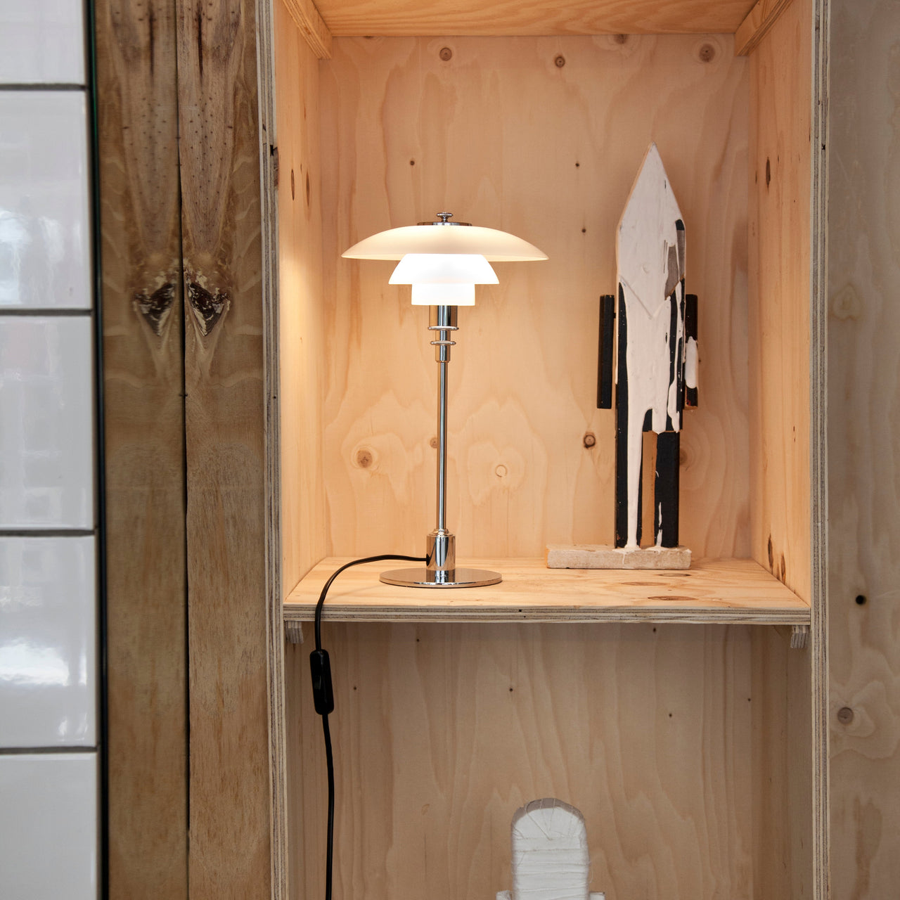 A table lamp with three white glass shades on a chrome stem, lit on a plywood shelf