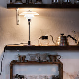 A glowing table lamp with layered white glass shades on a chrome stem, on a wooden shelf beside coffee kettles