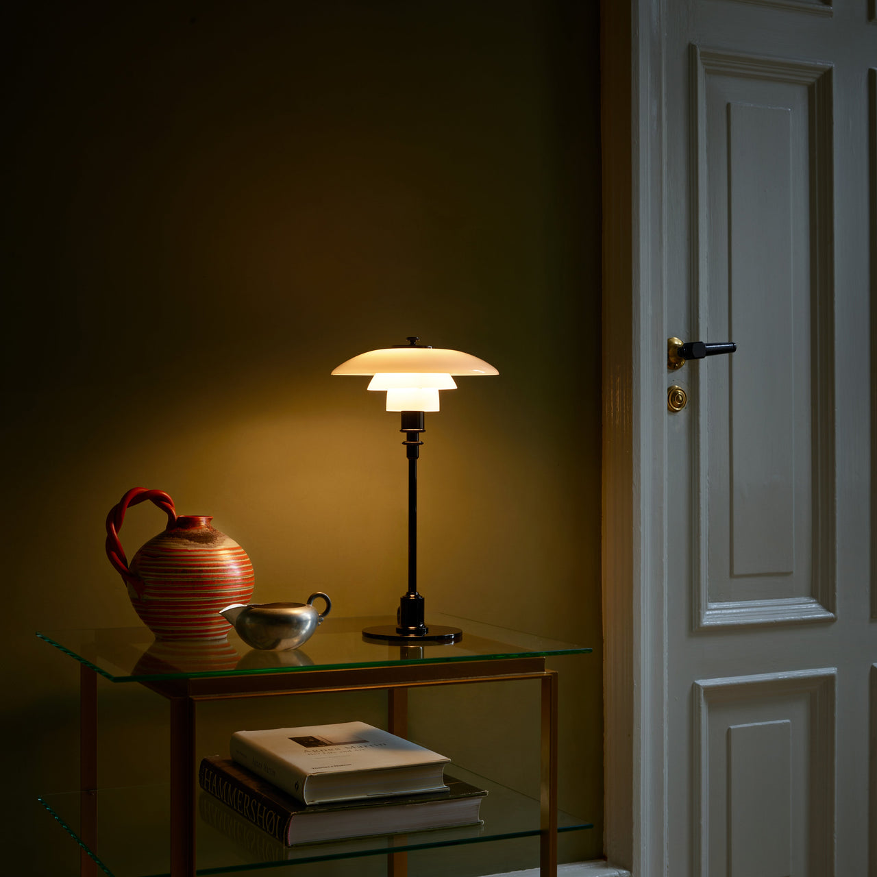 A glowing table lamp with three layered white glass shades on a slim black stem, on a glass side table by a white door
