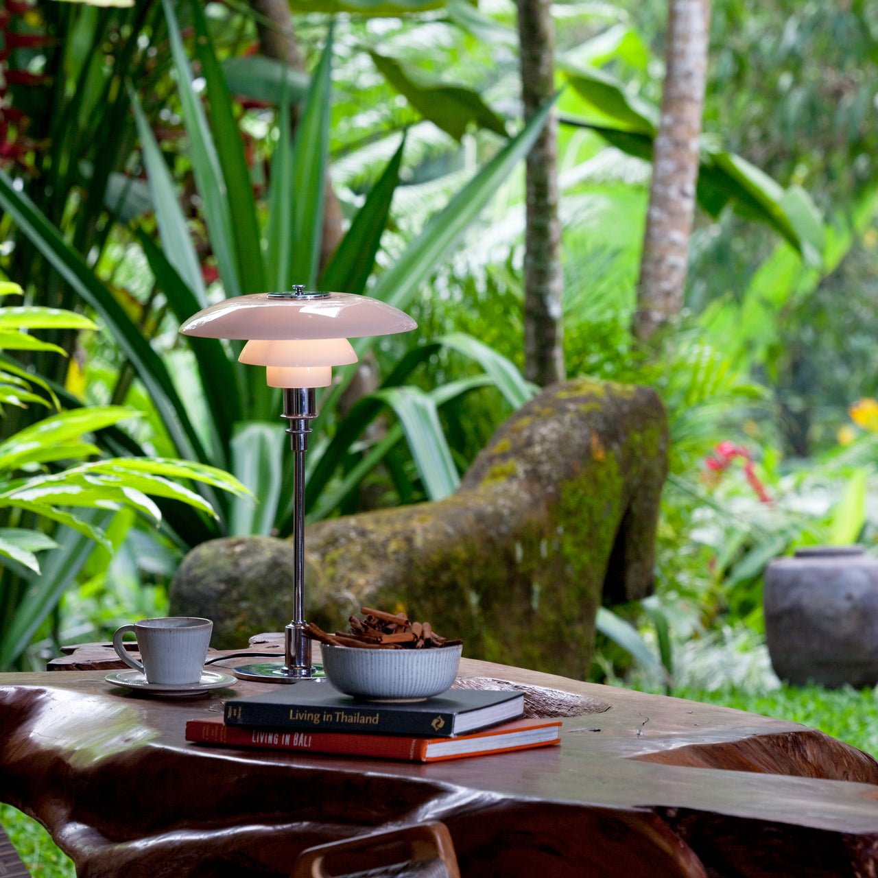 A table lamp with layered pale pink glass shades on a slim chrome stem, on a wooden table in a garden