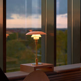 A lit brass table lamp with layered glass shades sits on a wooden block by a window at dusk