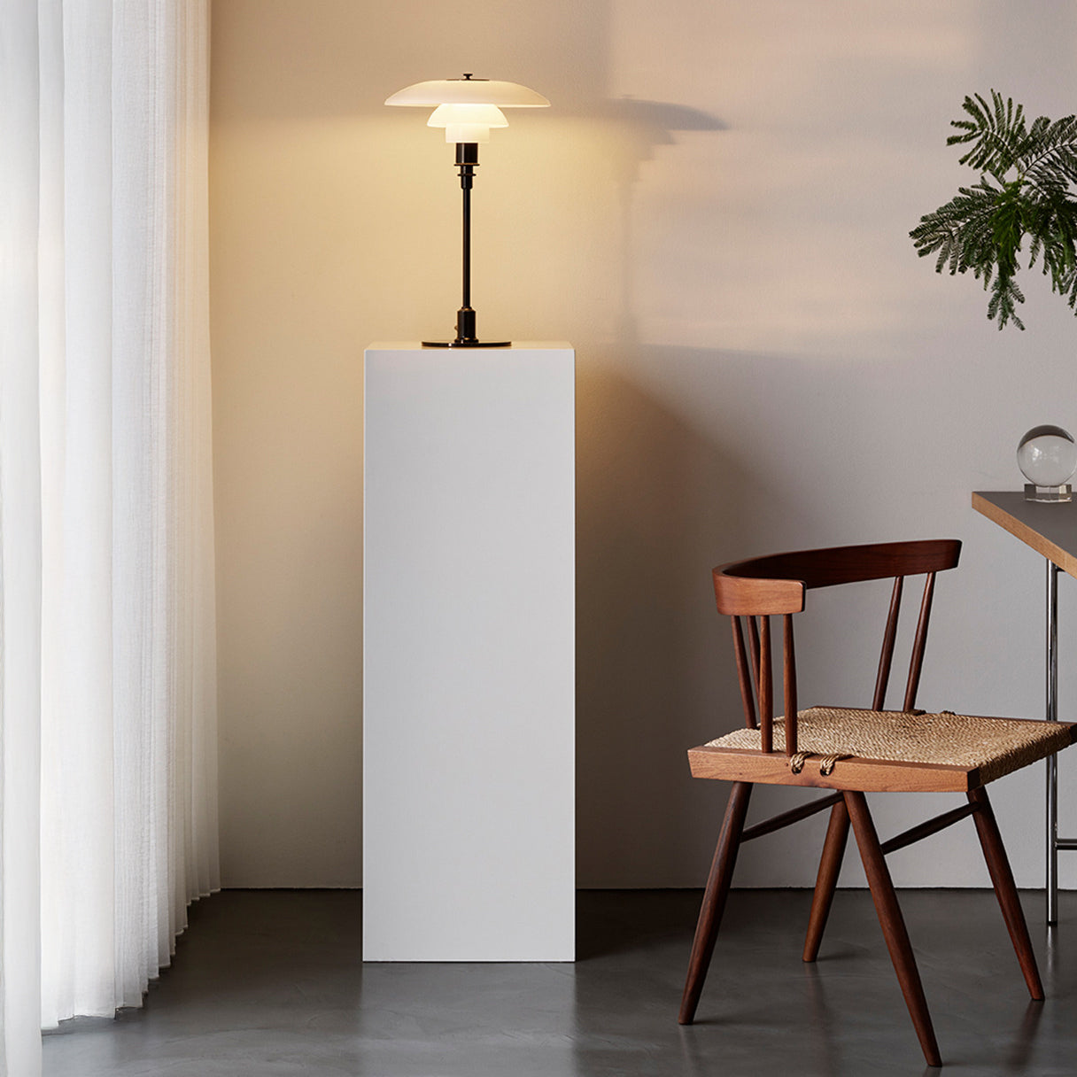 A lit table lamp with layered white glass shades on a white plinth beside a wooden spindle-back chair
