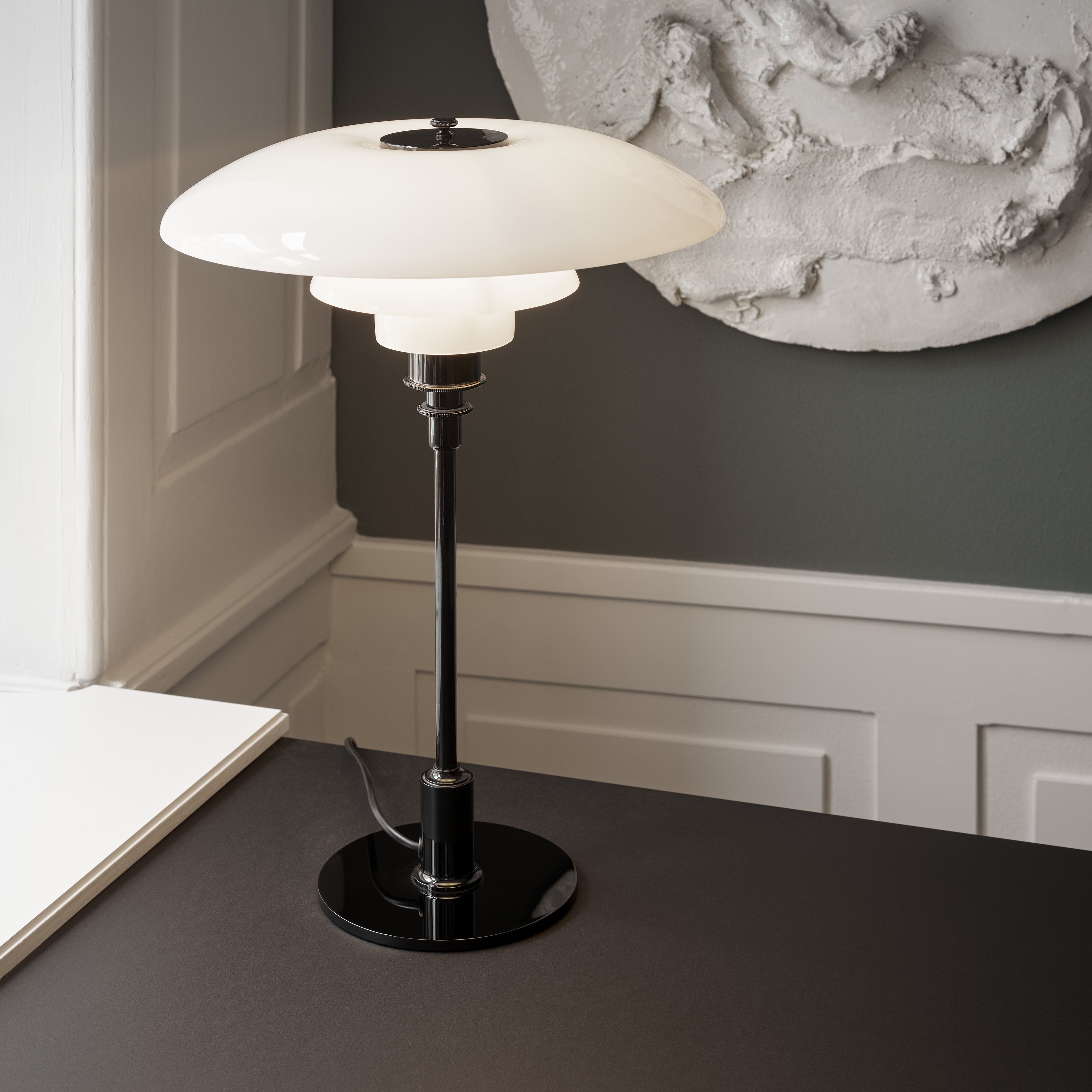 A table lamp with three layered white glass shades on a slim black stem and round base, on a dark desk