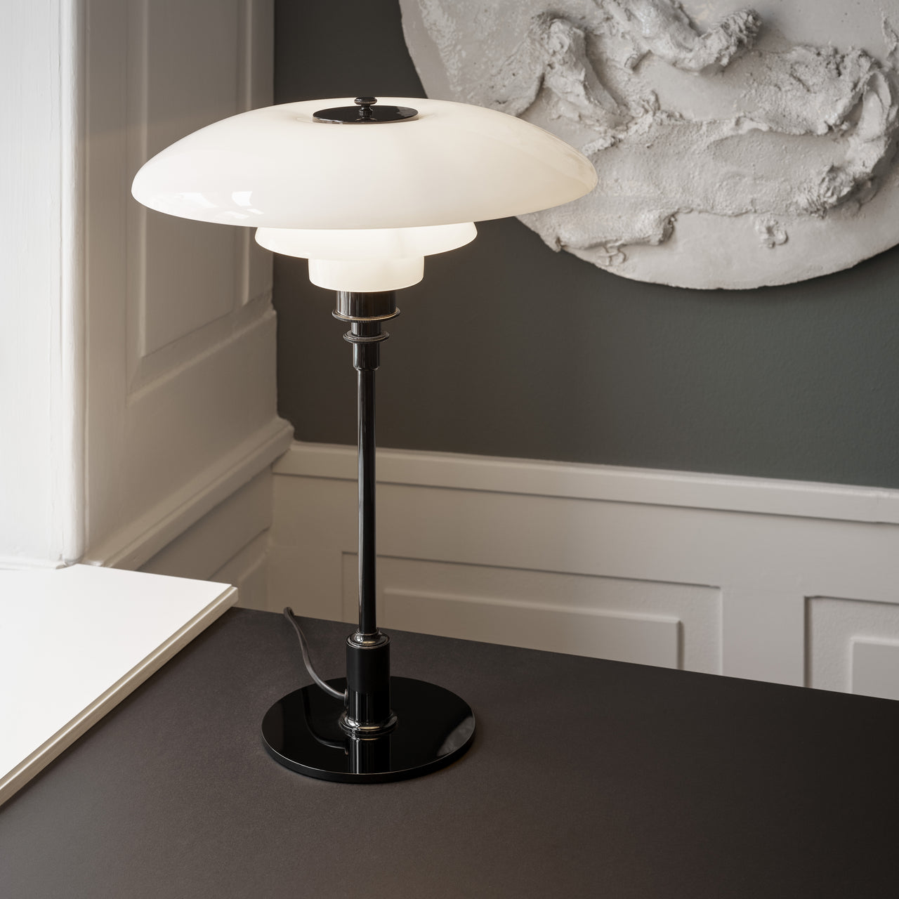 A table lamp with three layered white glass shades on a slim black stem and round base, on a dark desk