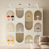 Mushroom-shaped table lamps in many colours on a cream shelving unit with arched niches, beside a beige sofa