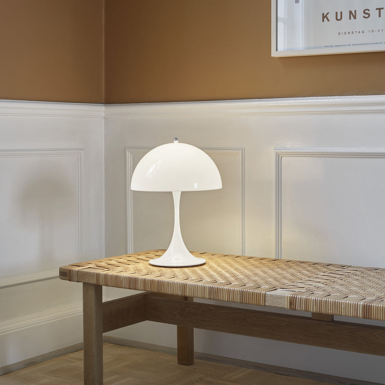 A glowing white mushroom-shaped table lamp on a woven cane and oak bench in a white-panelled hallway