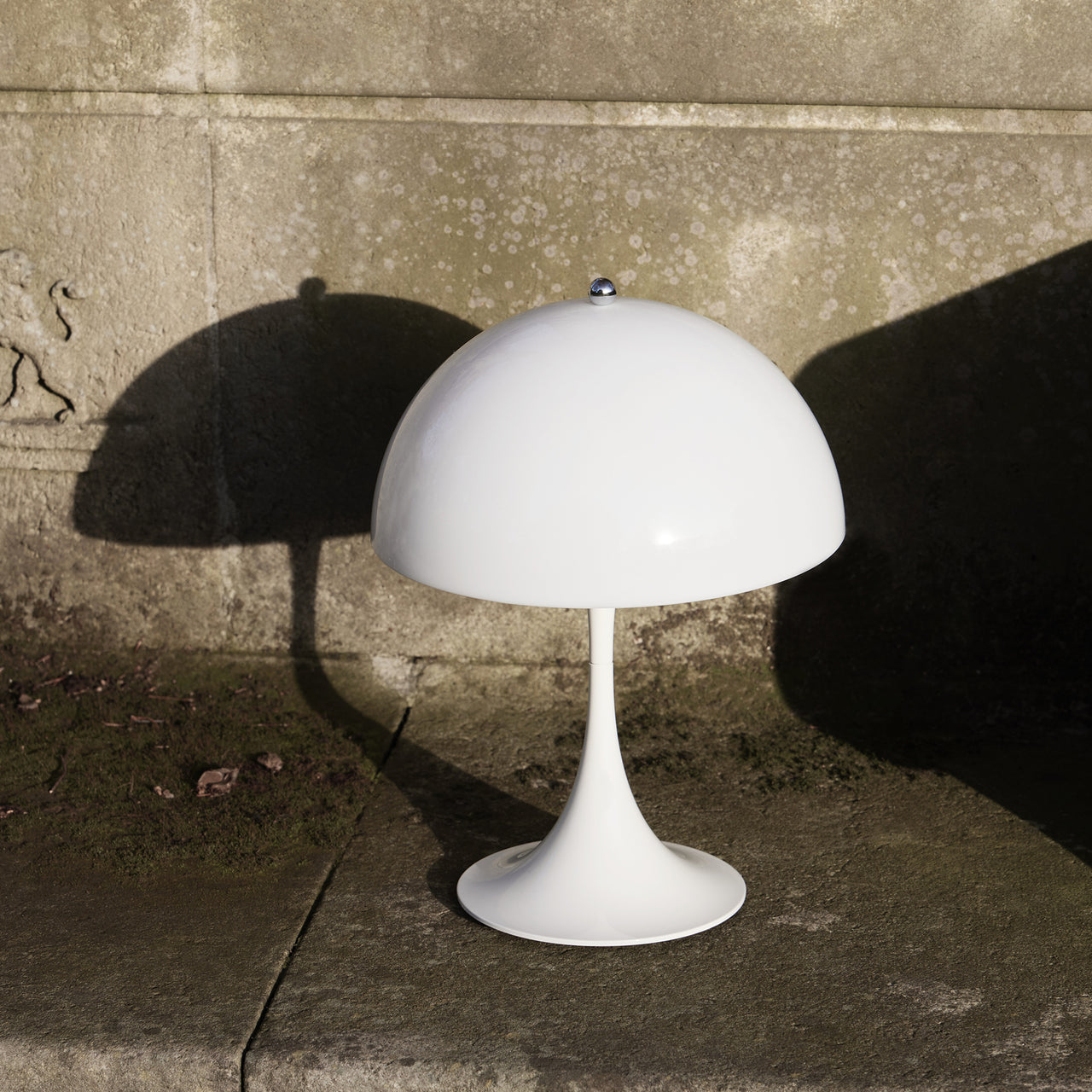 A white table lamp with a domed shade and flared base on a stone ledge, casting shadows on a weathered wall