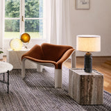 A square stone side table with a lamp on top, beside a rust-orange lounge chair in a sunlit room