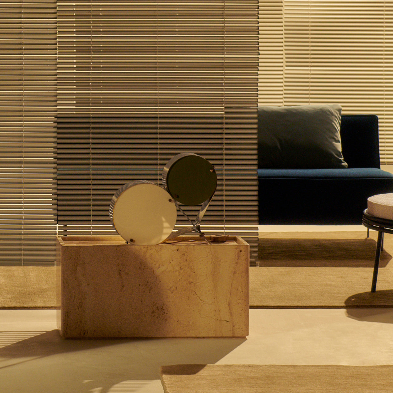 A rectangular travertine side table holding two round discs, beside a navy sofa and slatted blinds