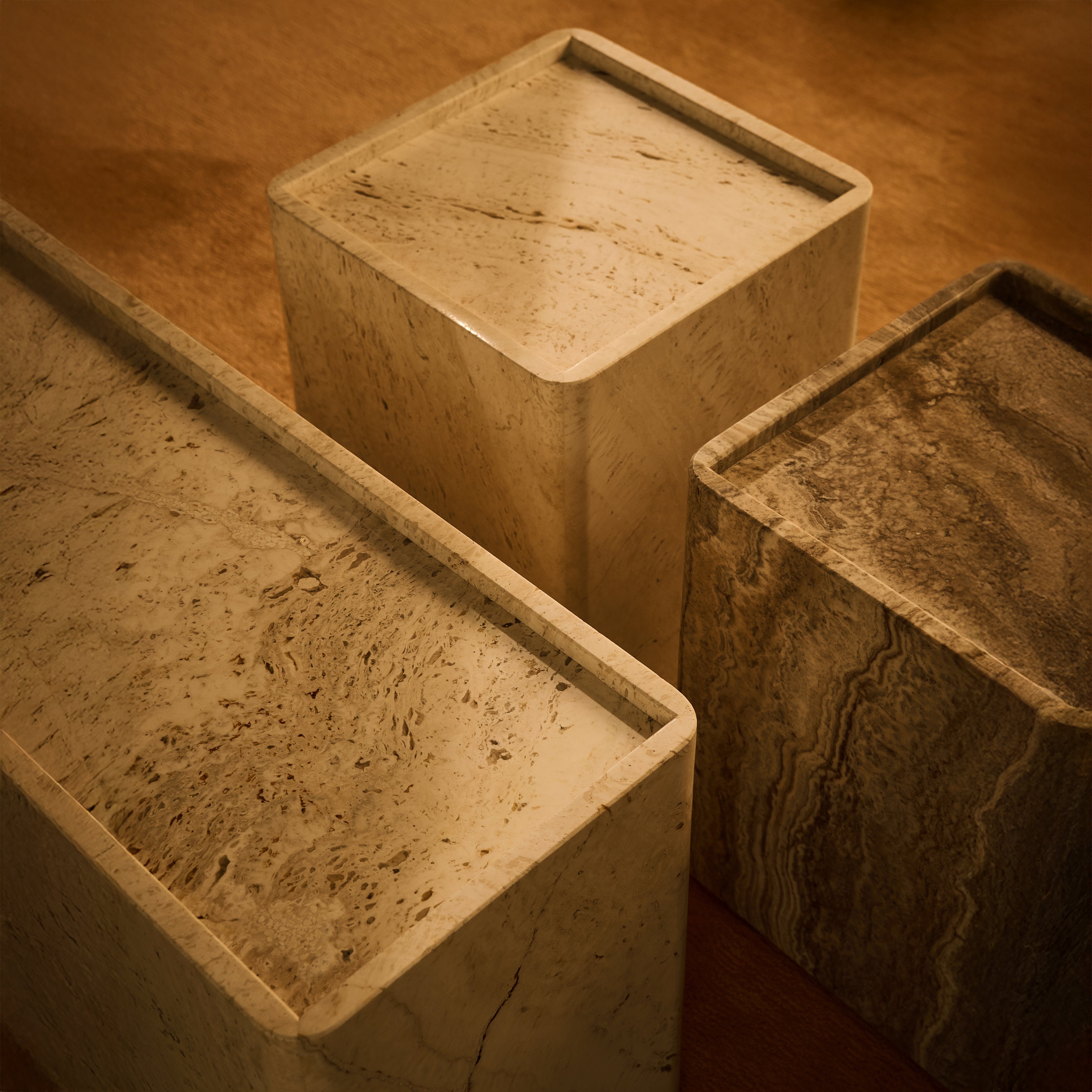 Overhead close-up of three square stone side tables in cream and grey-brown travertine with raised rims