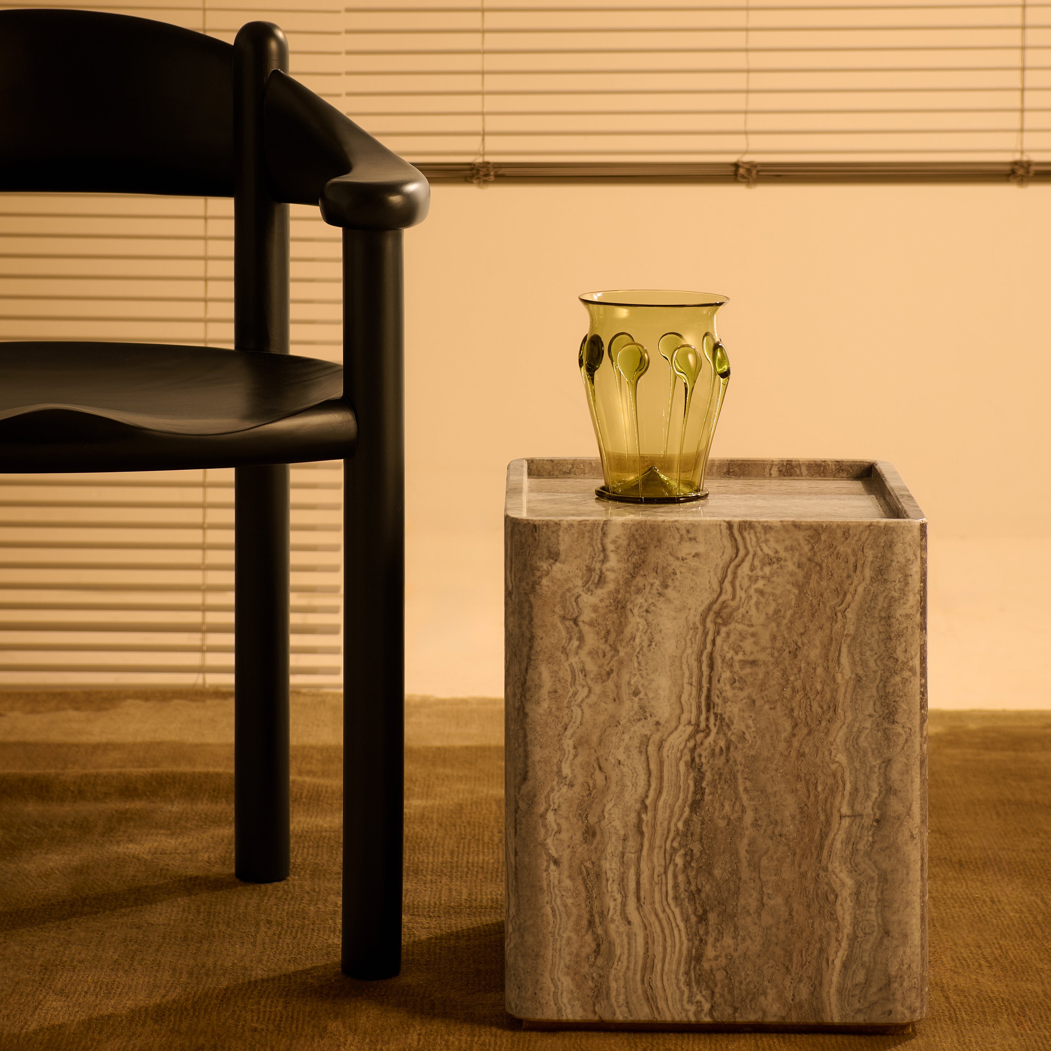 A square side table in banded grey-brown stone with an olive glass vase, beside a black wooden chair