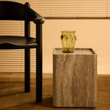 A square side table in banded grey-brown stone with an olive glass vase, beside a black wooden chair