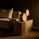 A low striped stone side table holding a brass twin-disc lamp, beside a cream sofa in a dim room