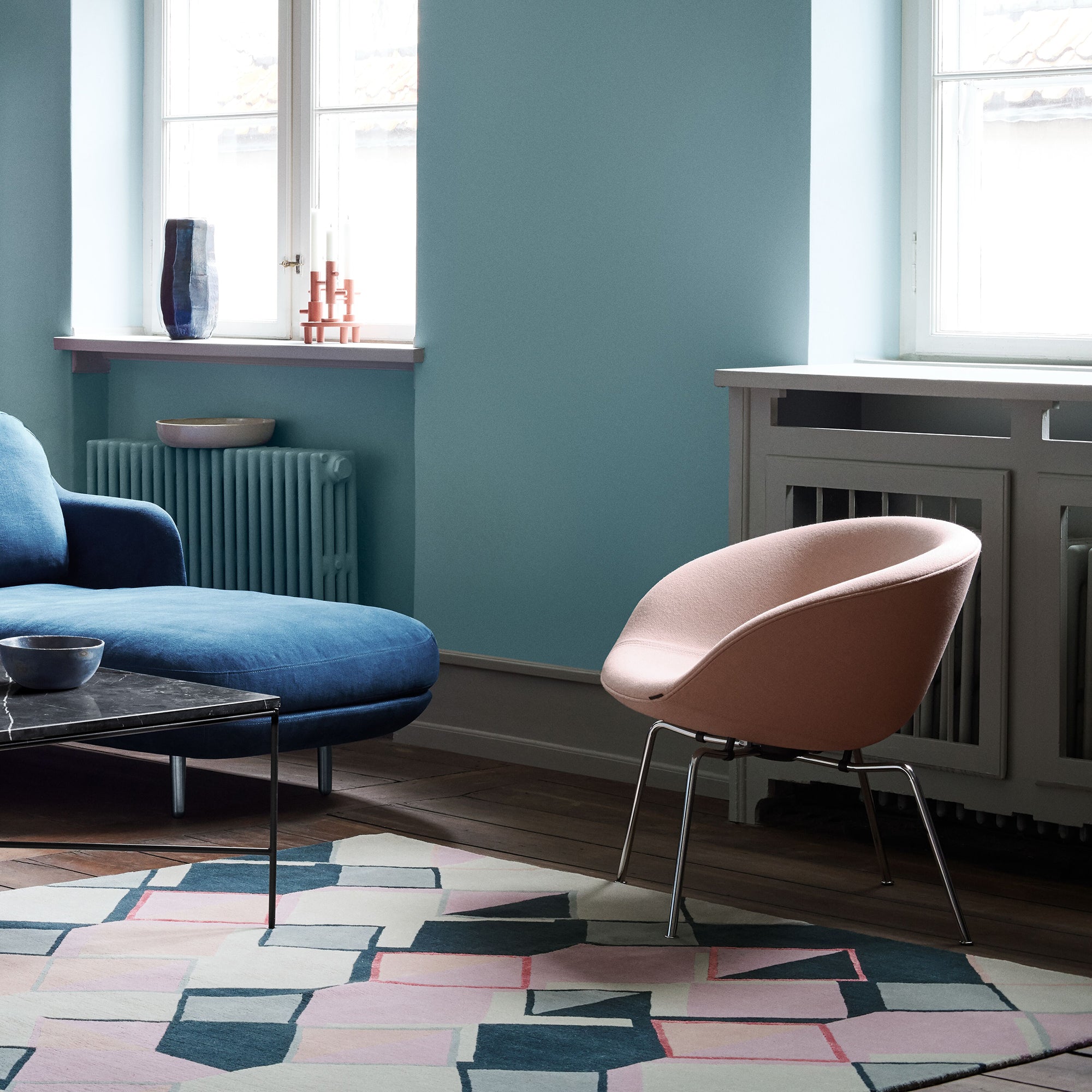 A rounded pale pink lounge chair on slim chrome legs in a teal room with a blue sofa