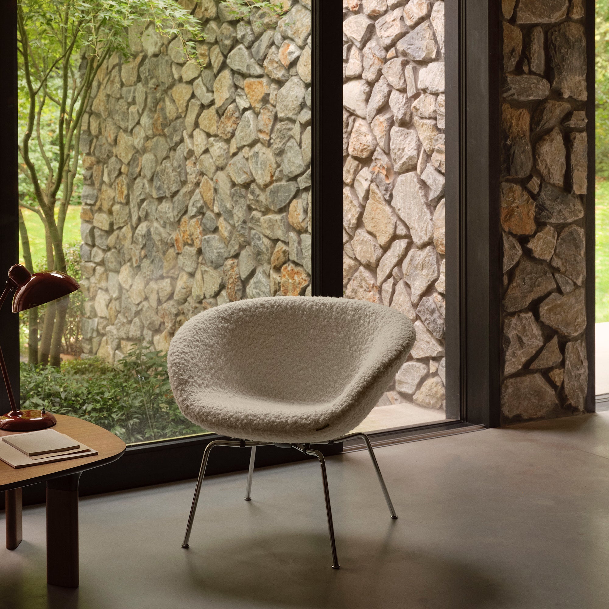 A rounded lounge chair in cream bouclé fabric on slim chrome legs beside a stone-walled window