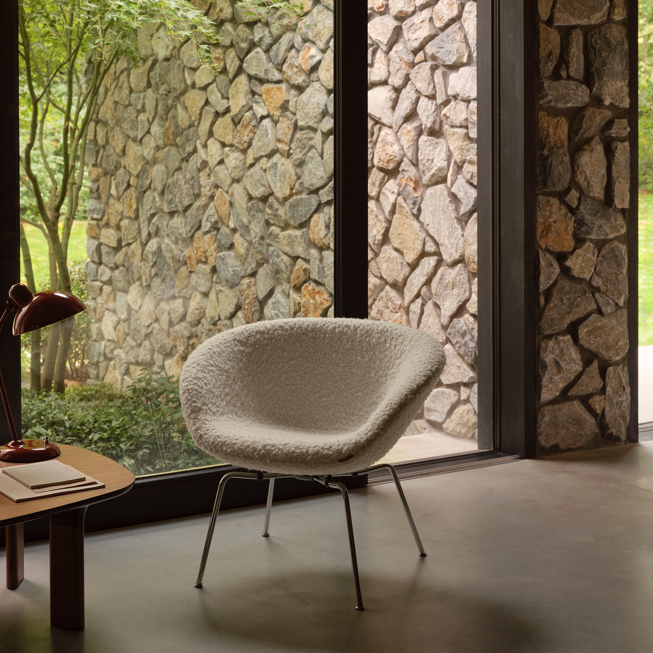 A rounded lounge chair in cream bouclé fabric on slim chrome legs beside a stone-walled window