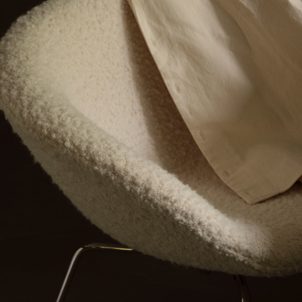Close-up of a curved cream bouclé chair shell on thin chrome legs with a cloth draped over it