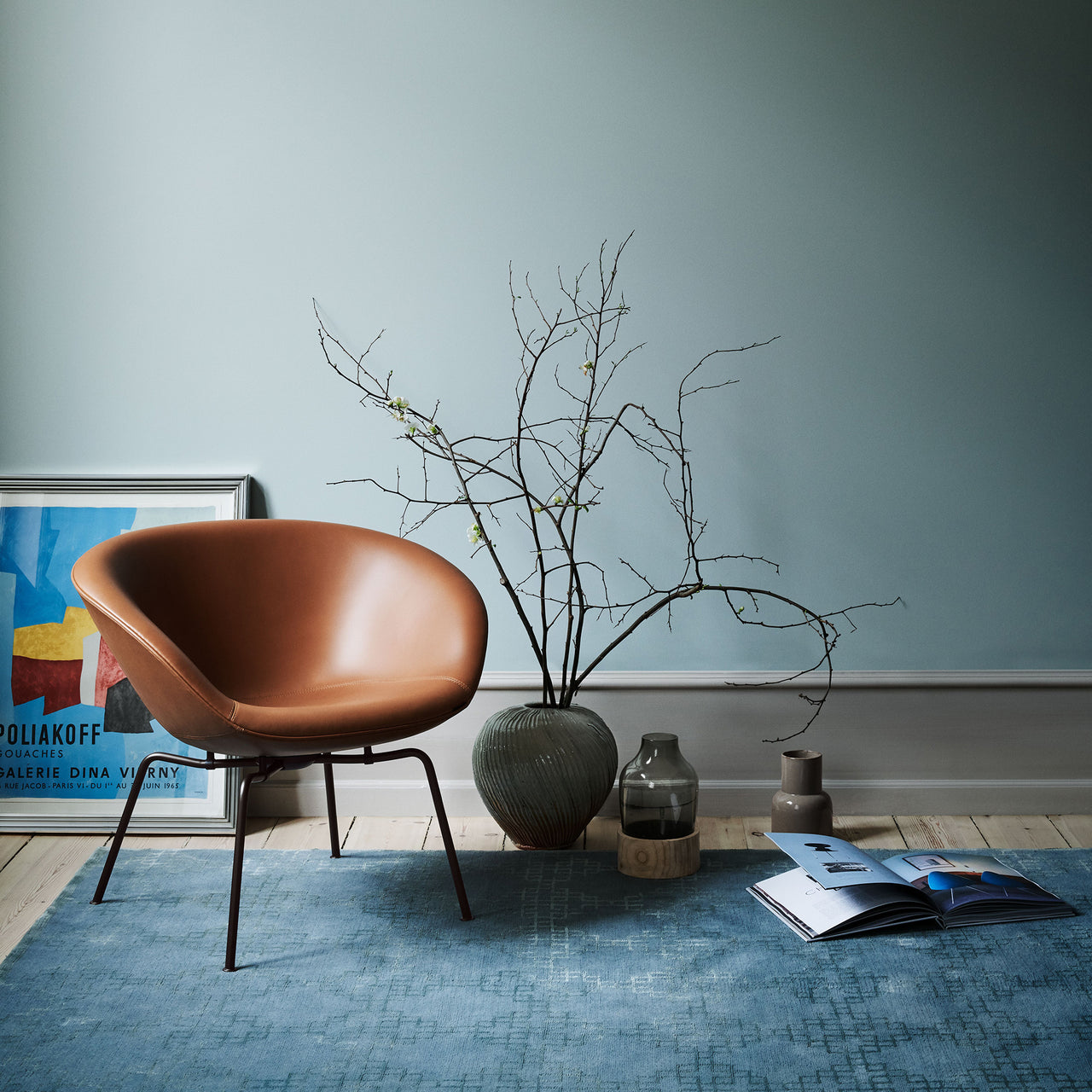 A rounded tan leather lounge chair on thin dark legs, on a blue rug by a pale blue wall