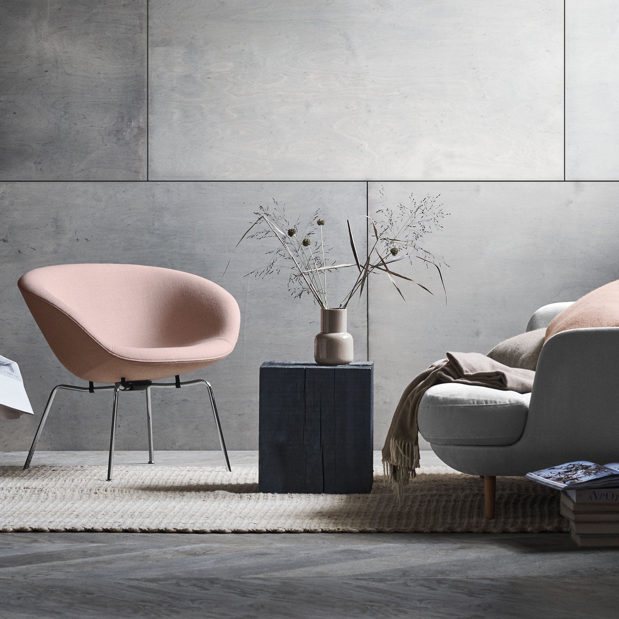 A pale pink bowl-shaped lounge chair on slim chrome legs beside a dark block table and a grey sofa