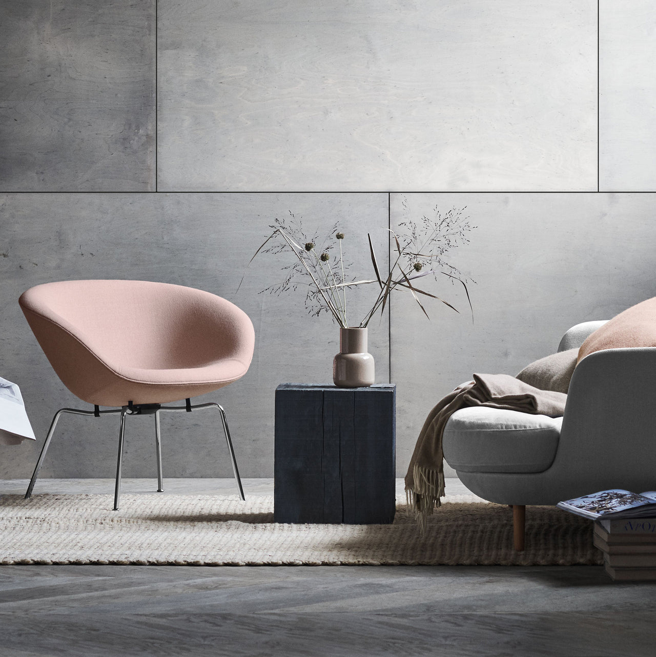 A pale pink bowl-shaped lounge chair on slim chrome legs beside a dark block table and a grey sofa