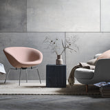 A pale pink bowl-shaped lounge chair on slim chrome legs beside a dark block table and a grey sofa