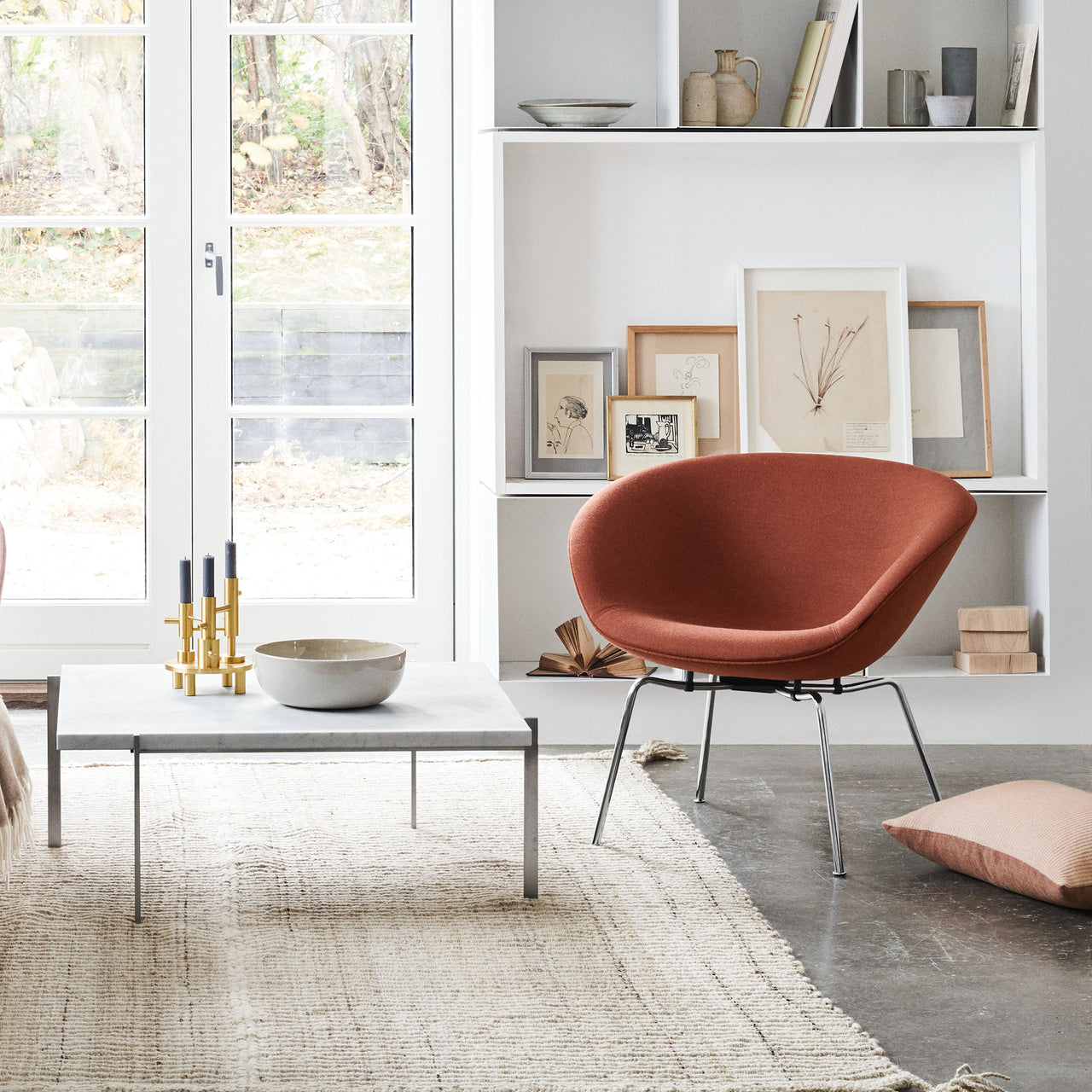 A rust-orange bowl-shaped lounge chair on slim chrome legs beside a marble coffee table by a white bookcase