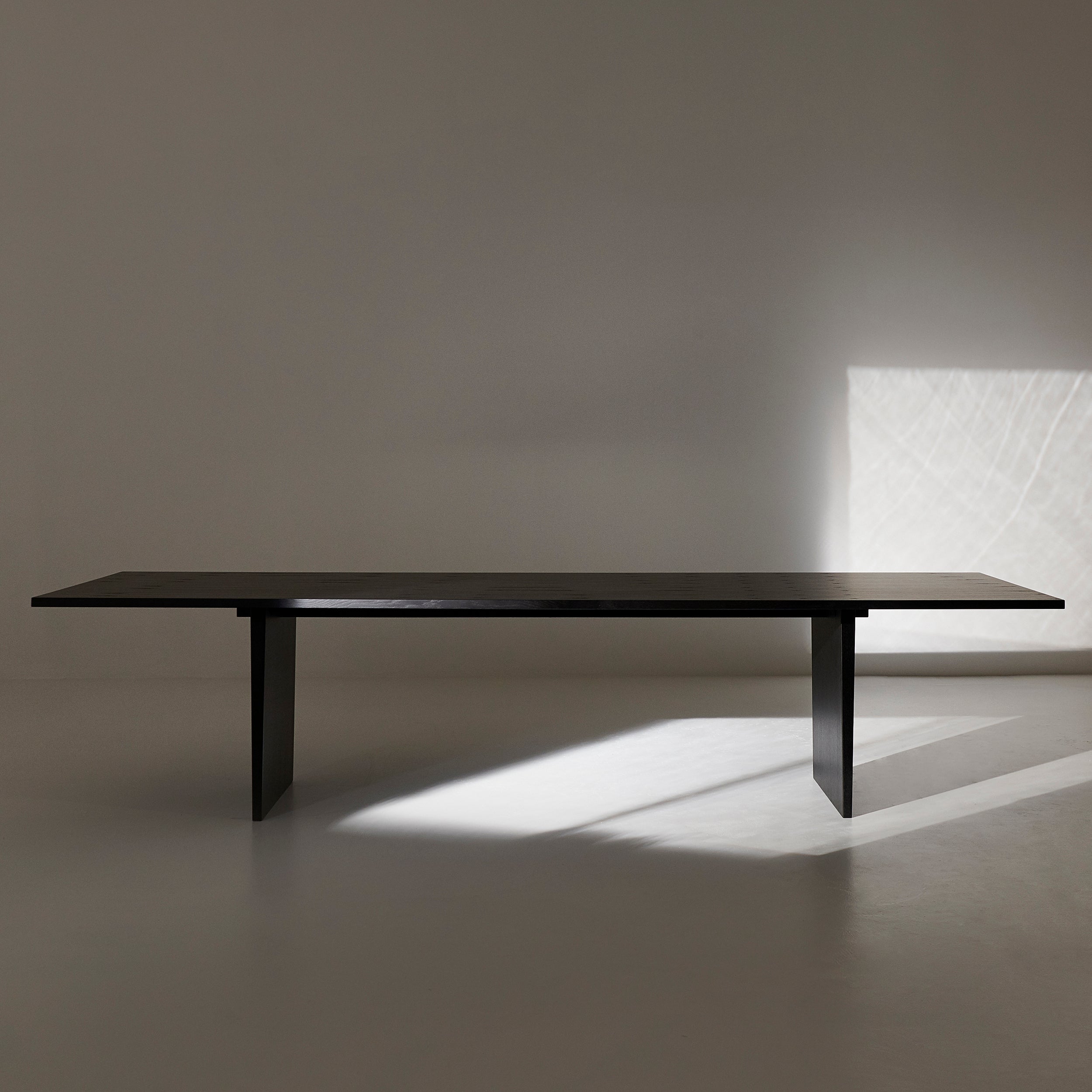 A long black wooden table on two slab legs in a bare room with sunlight on the floor