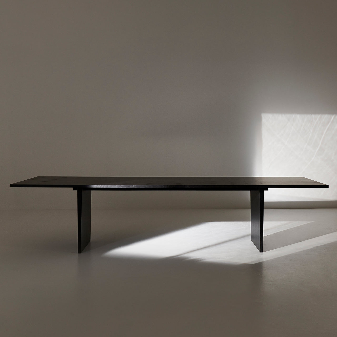 A long black wooden table on two slab legs in a bare room with sunlight on the floor