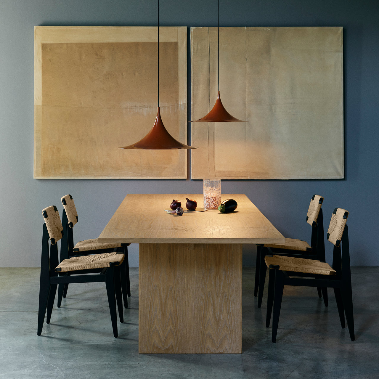 A light oak dining table with a slab base and black chairs with woven seats under two rust pendants