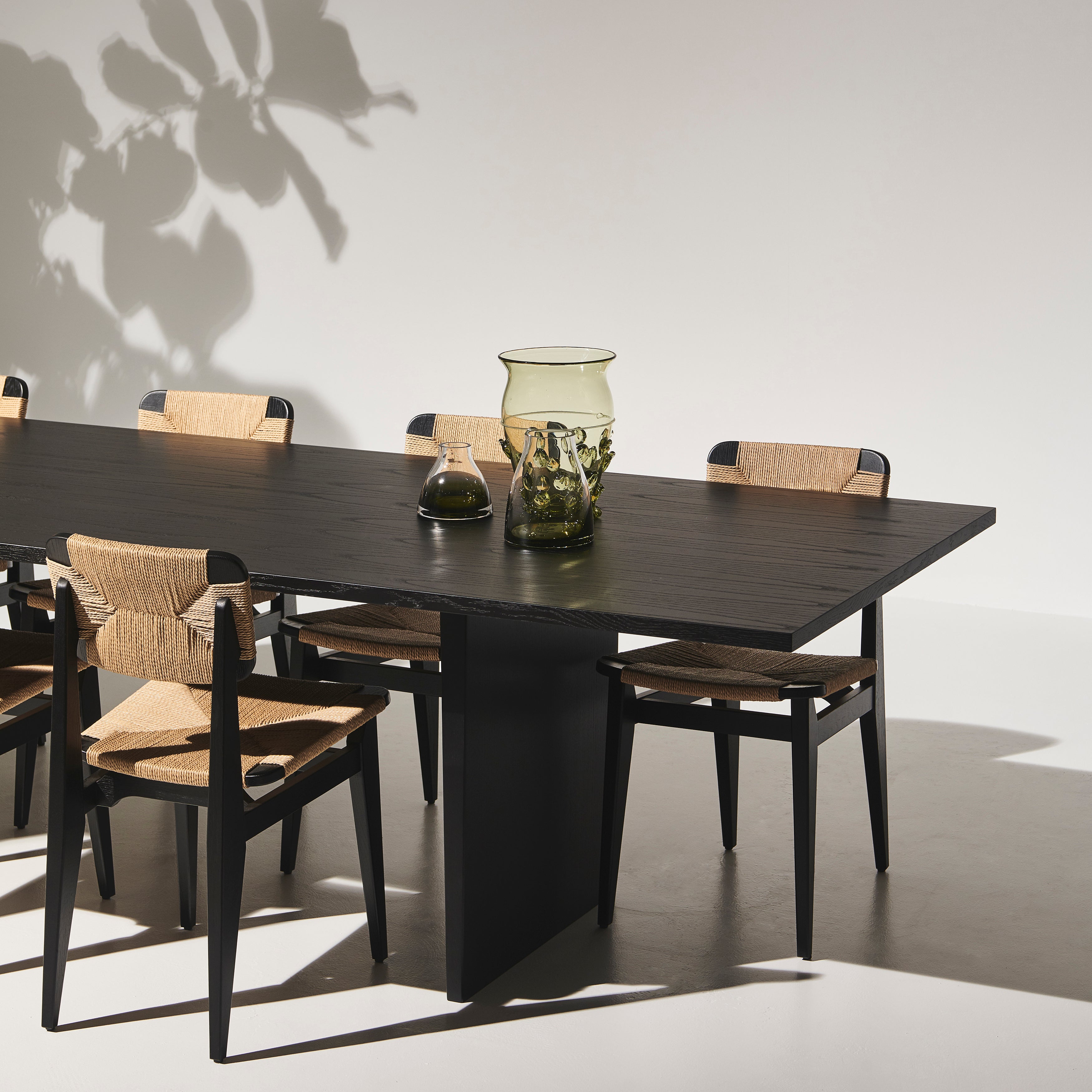 A black wood dining table with a slab base, surrounded by black chairs with woven natural seats
