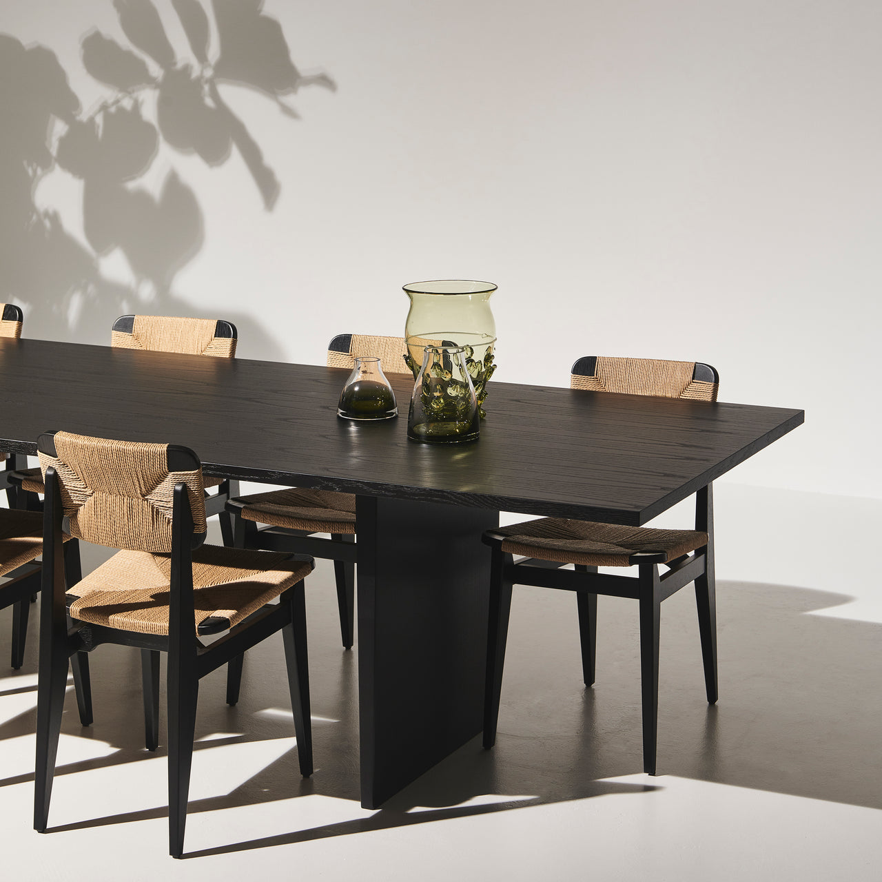 A black wood dining table with a slab base, surrounded by black chairs with woven natural seats