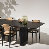 A black wood dining table with a slab base, surrounded by black chairs with woven natural seats