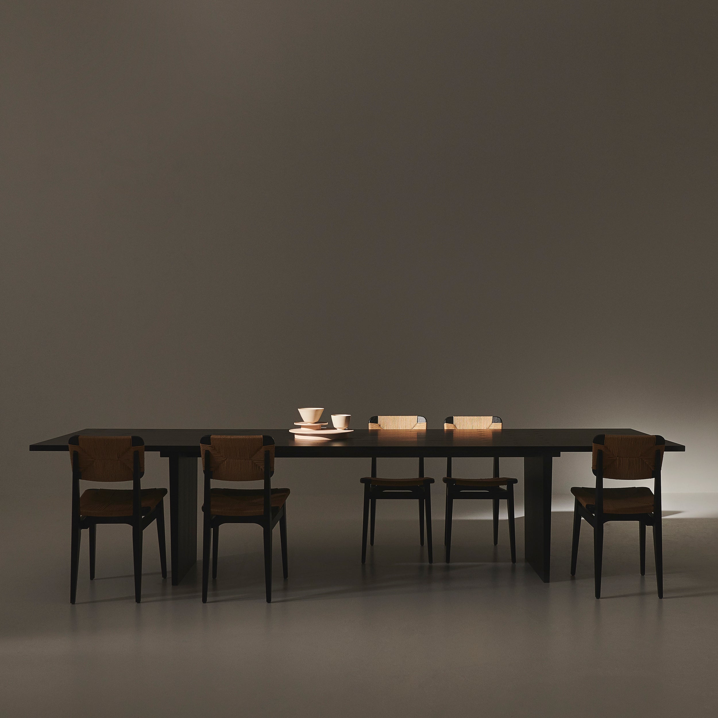 A long dark rectangular table on slab legs with five black chairs with woven backs in a dim room