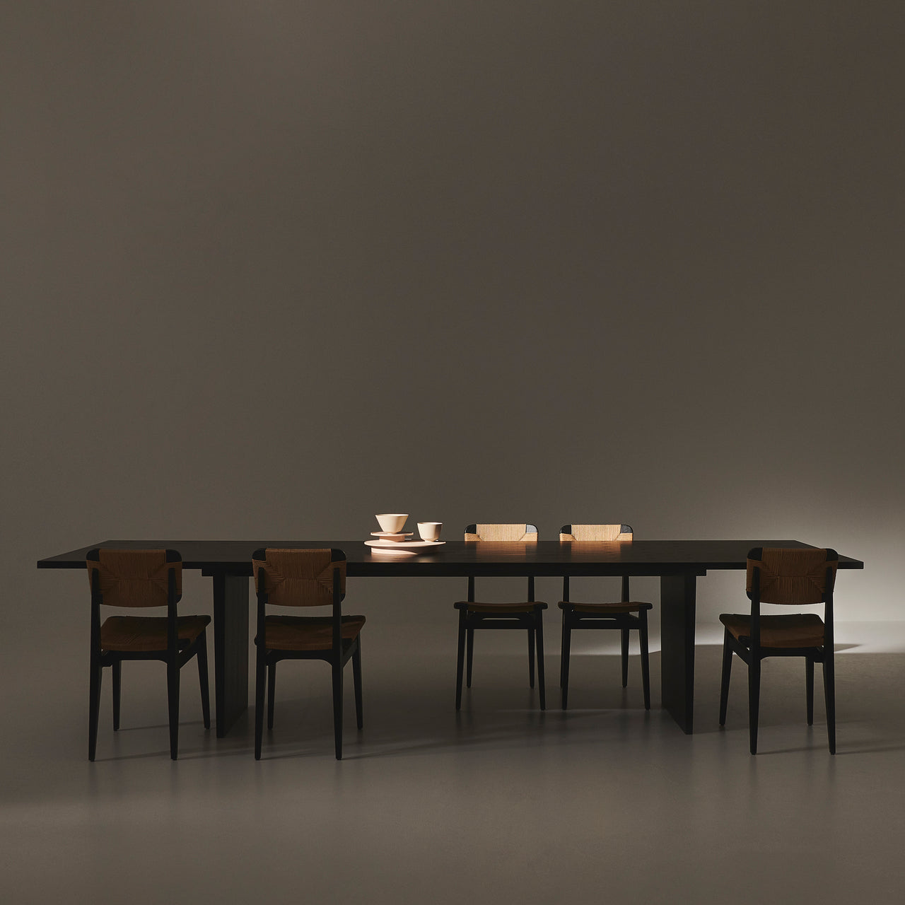 A long dark rectangular table on slab legs with five black chairs with woven backs in a dim room