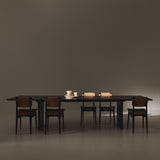 A long dark rectangular table on slab legs with five black chairs with woven backs in a dim room