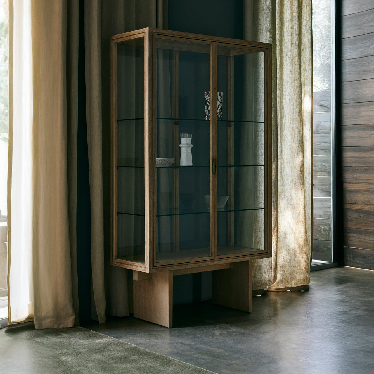 A tall oak-framed glass display cabinet with two doors and glass shelves, standing in a room with linen curtains