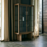 A tall oak-framed glass display cabinet with two doors and glass shelves, standing in a room with linen curtains