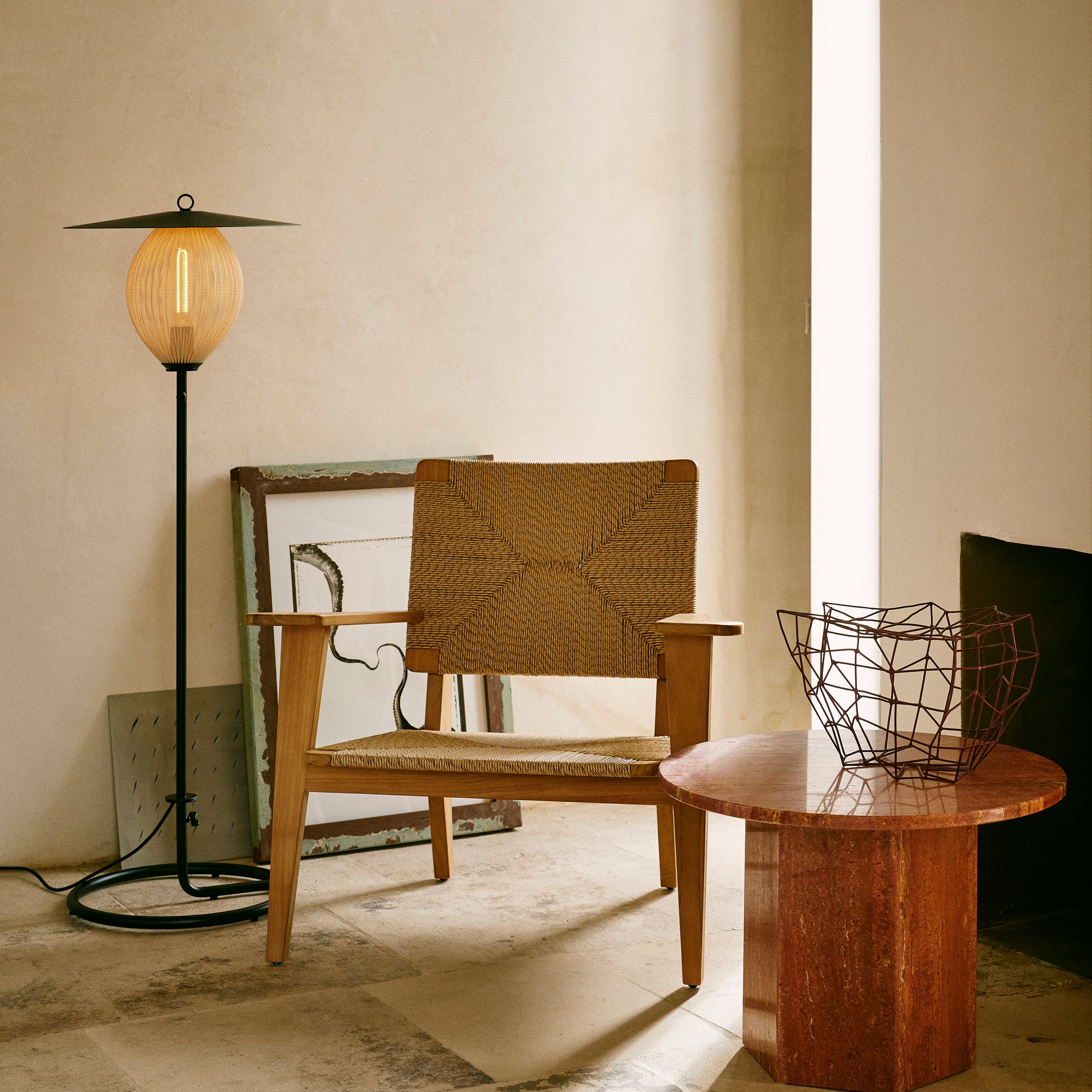 A black floor lamp with a round glowing amber shade beside a woven wooden armchair and a red stone side table