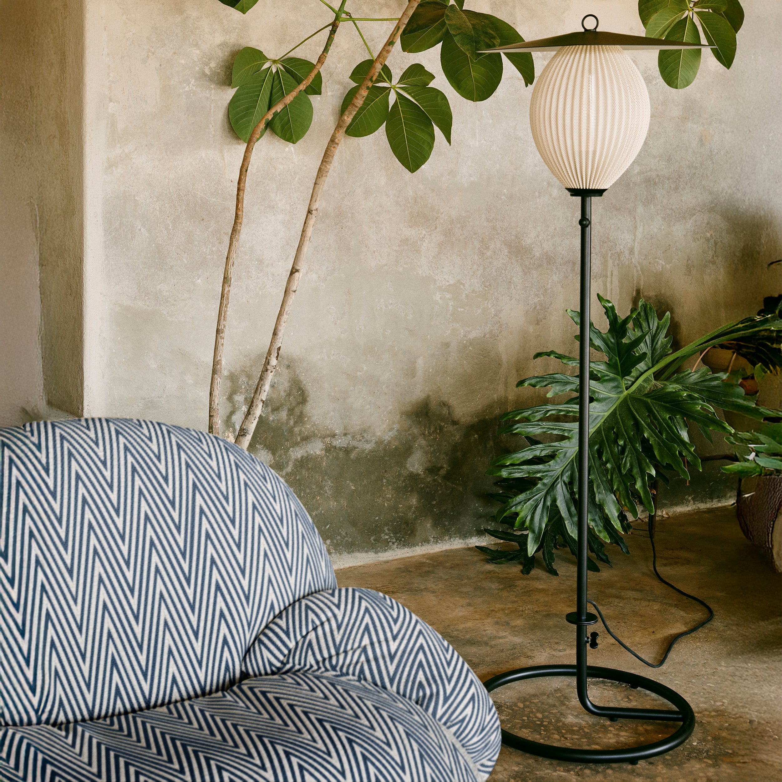 A black floor lamp with a round ribbed white shade and flat metal top beside green plants and a chevron-patterned chair