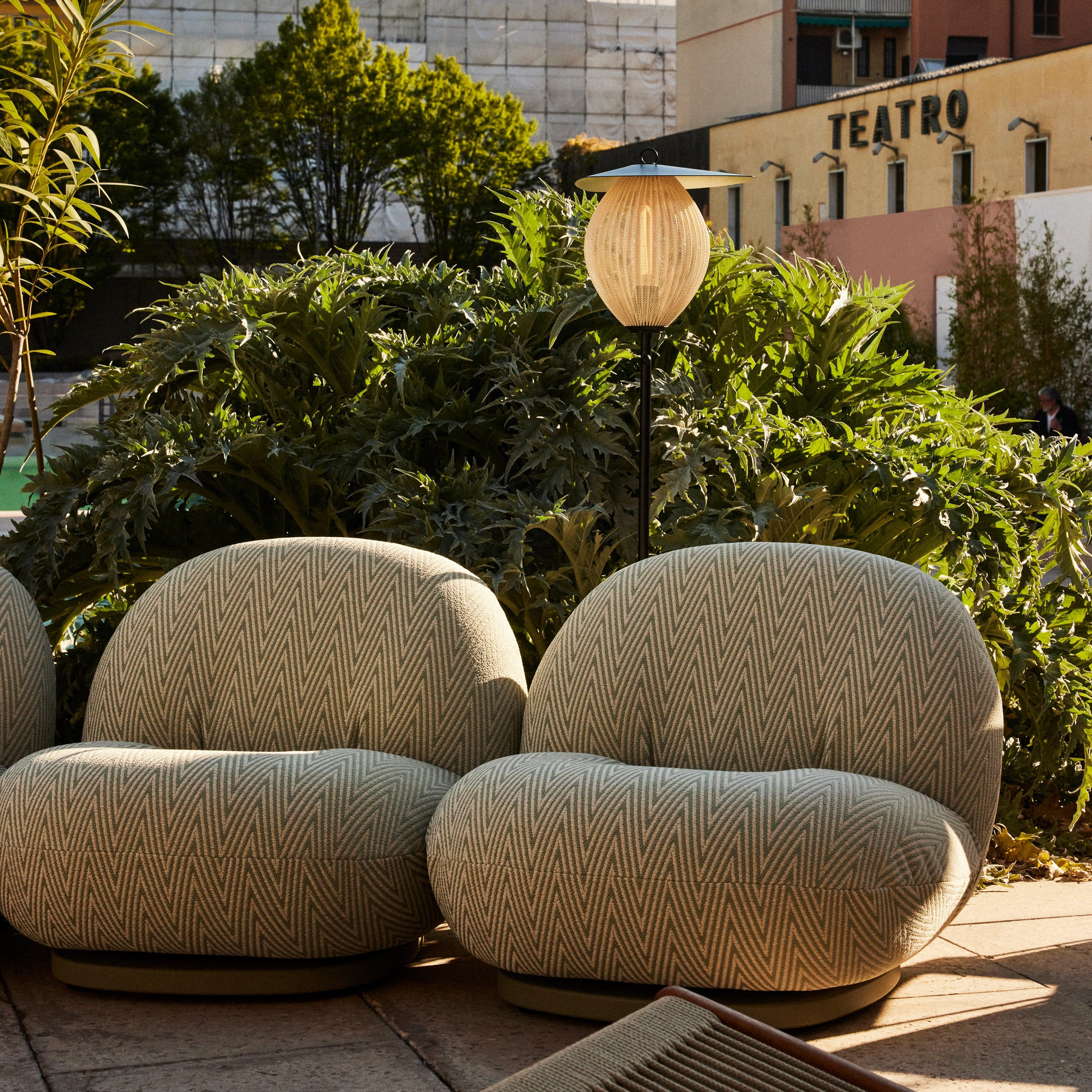 A slim outdoor floor lamp with a ribbed oval shade stands between two rounded green chevron armchairs