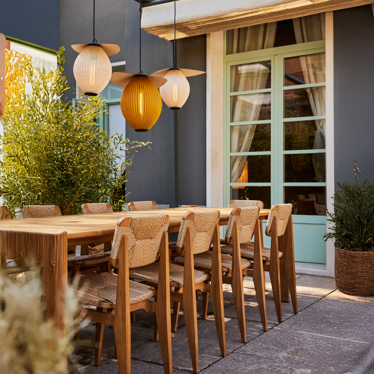 Three pleated pendant lights, one amber, above a wooden table with woven-seat chairs on a patio