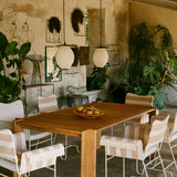 Two pale ribbed globe pendants hang over a teak table with striped chairs in a plant-filled courtyard
