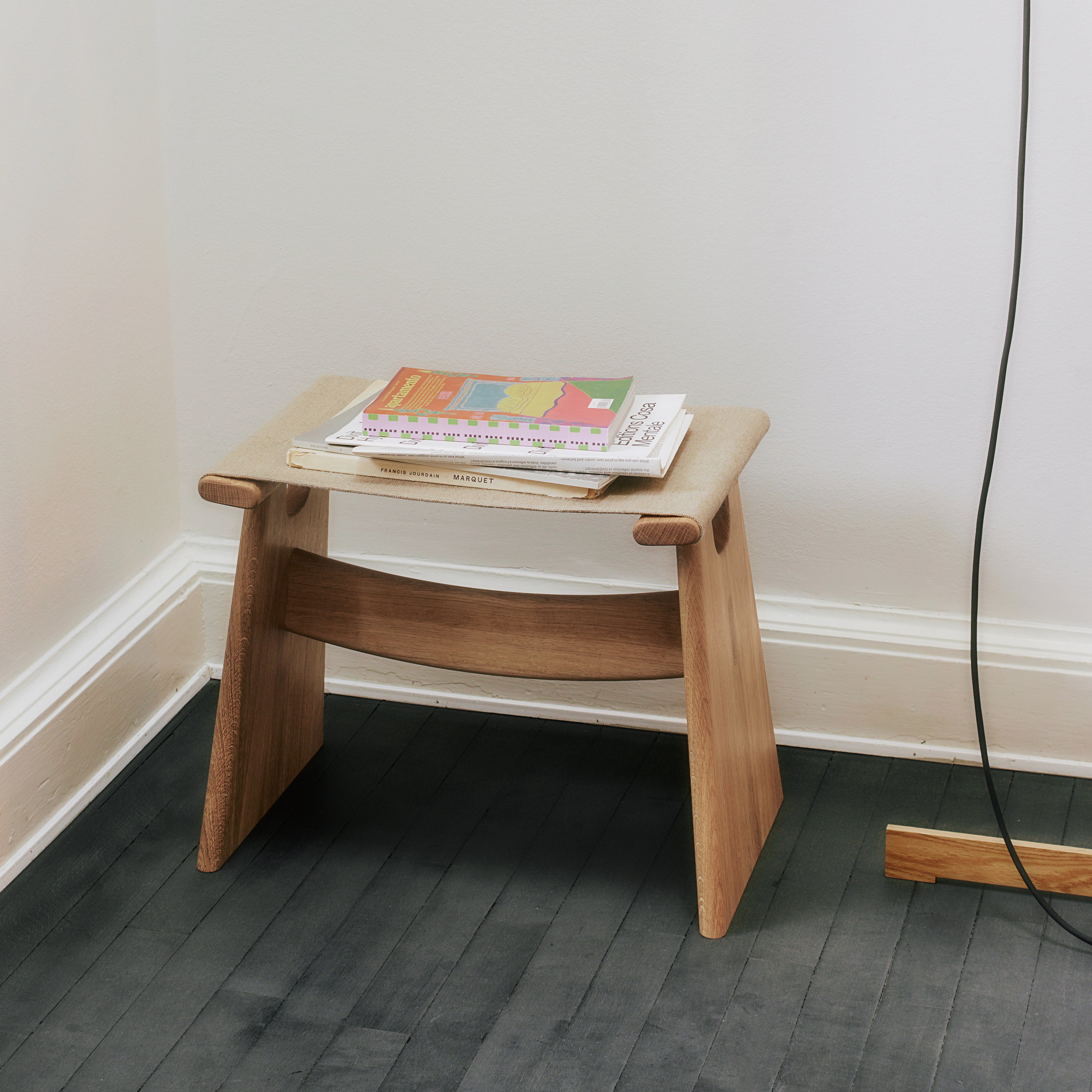 A low light oak stool with a beige felt seat and two slanted legs joined by a curved rail, on a dark floor