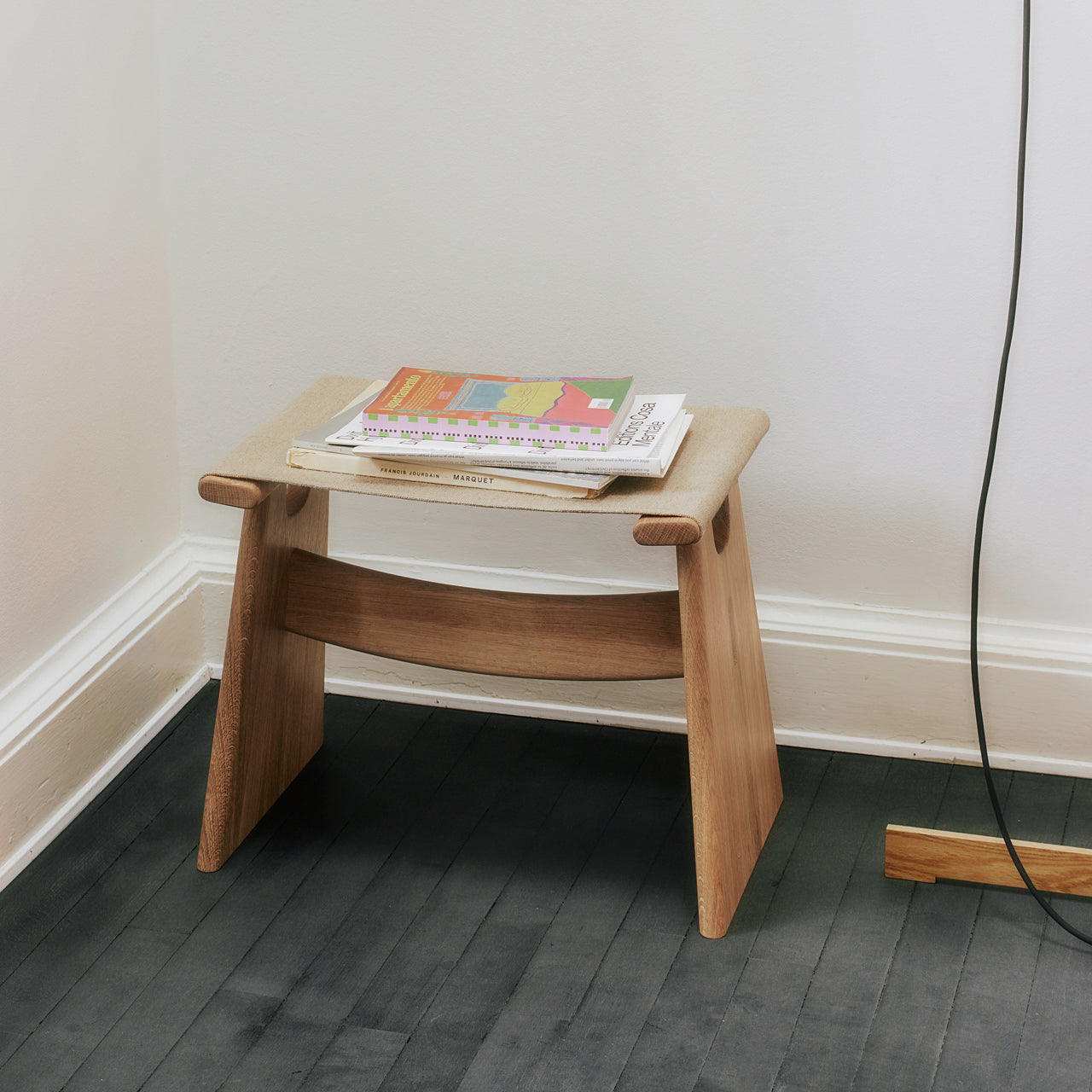 A low light oak stool with a beige felt seat and two slanted legs joined by a curved rail, on a dark floor