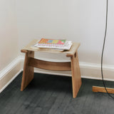 A low light oak stool with a beige felt seat and two slanted legs joined by a curved rail, on a dark floor