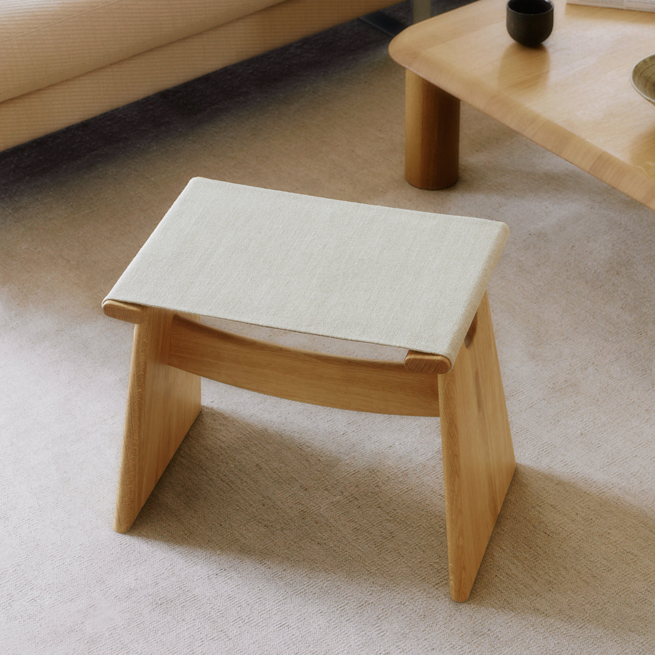 A low light oak stool with angled slab legs and a cream fabric sling seat on a pale rug