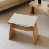 A low light oak stool with angled slab legs and a cream fabric sling seat on a pale rug