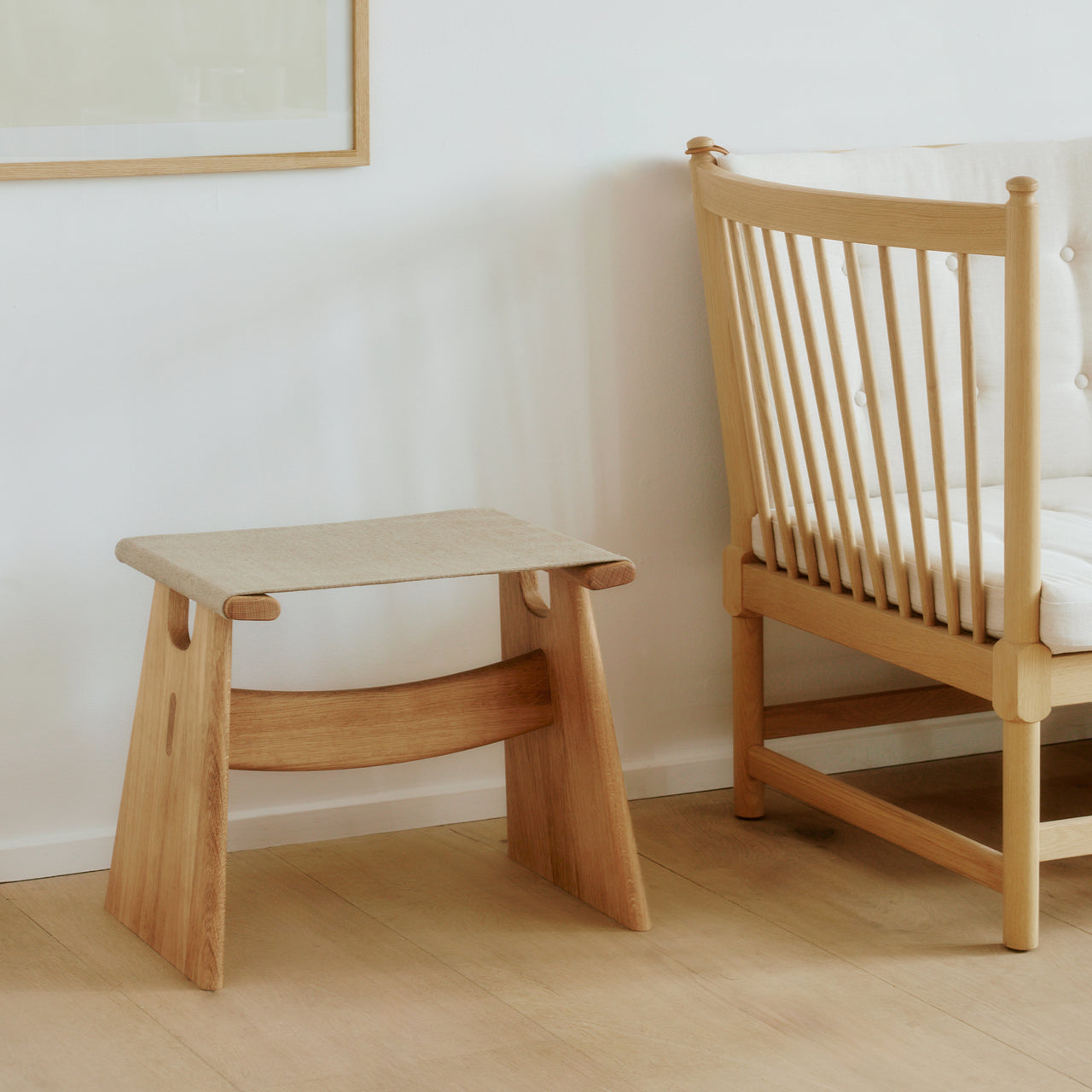 A light oak stool with a beige fabric seat and curved crossbar, beside a spindle-back chair