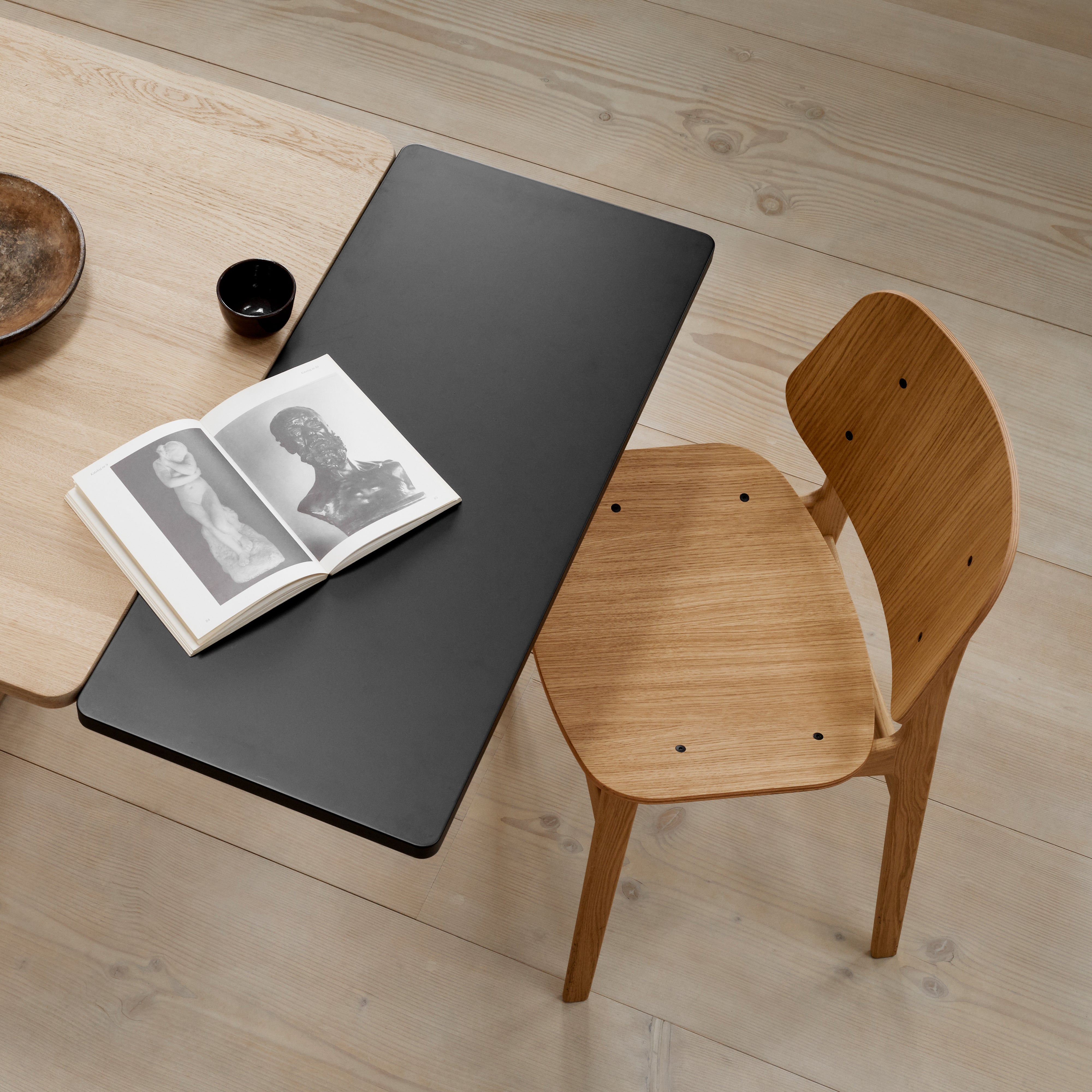 Overhead view of a light oak chair with a curved back beside a black rectangular tabletop on a wide plank floor