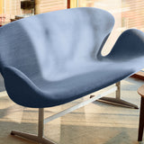 A curved two-seat sofa in blue-grey fabric on a slim metal base, in a sunlit room